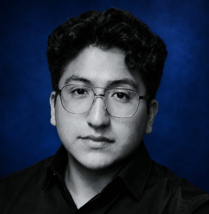 Portrait of a young man with round glasses and dark hair, wearing a black shirt, against a deep blue studio backdrop.