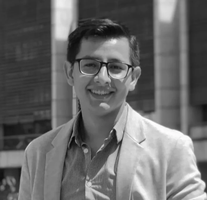 Young man with short dark hair and glasses smiling at the camera, wearing a light blazer and striped shirt outdoors.